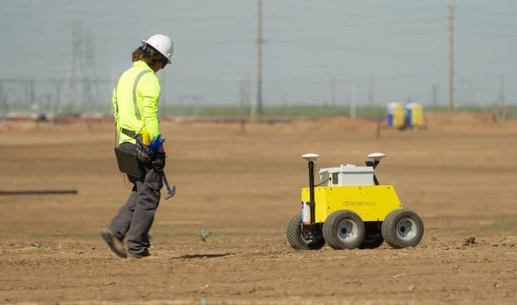 Crew member walking beside CivDot robotic survey equipment on a solar farm layout.